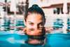 Budapest Thermen: Eine Frau ist bis knapp unter ihrer Nasenspitze im blauen Wasser eines Outdoor-Schwimmbeckens. Das Wasser hinter ihr glitzert im Sonnenlicht. 