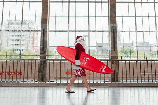 Der Weihnachtsmann mit rotem Surfbrett in der Haupthalle des Westbahnhofs spazierend.