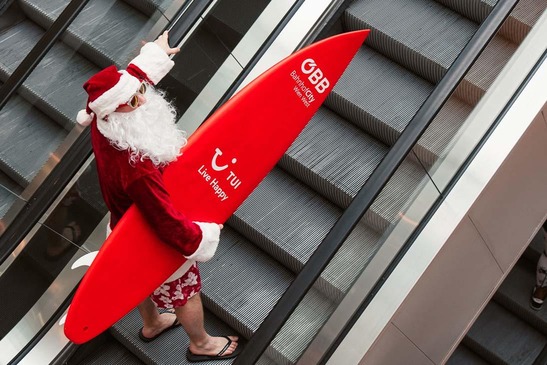 © ÖBB | Lukas Leonte Der Weihnachtsmann mit Surfbrett auf der Rolltreppe.