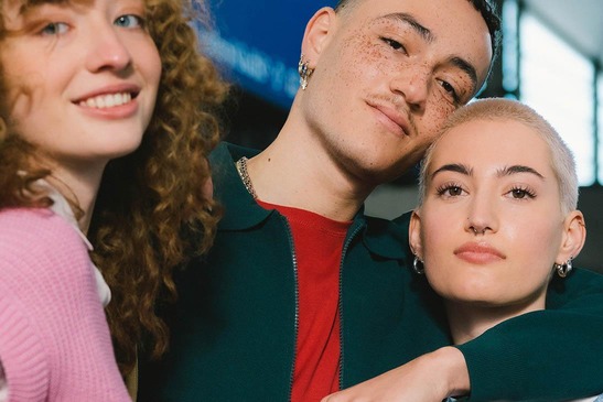 Zwei Frauen und ein Mann stehen auf dem Bahnsteig und lächeln in die Kamera. Der Mann legt seinen Arm um eine der Frauen. 