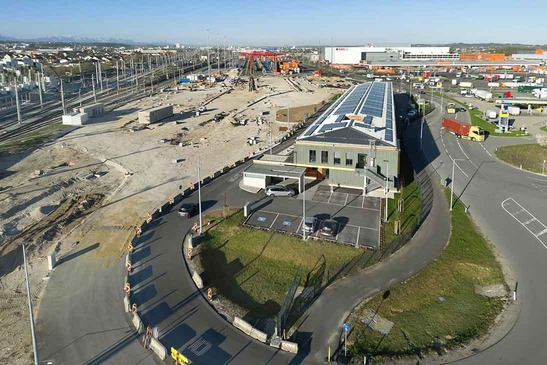 Luftaufnahme eines großen Terminalareals mit Baustelle, mehreren Gleisen, einem modernen Gebäude mit Solarpaneelen am Dach sowie umliegenden Straßen und Industrieflächen.