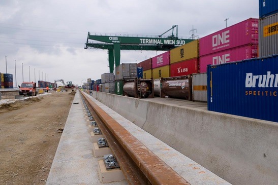 View along a newly built railway track in a container terminal Wien Süd with stacked freight containers and a green gantry crane in the background. 
