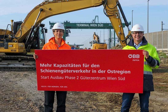 Two people are holding a sign, with an excavator and a gantry crane visible in the background.