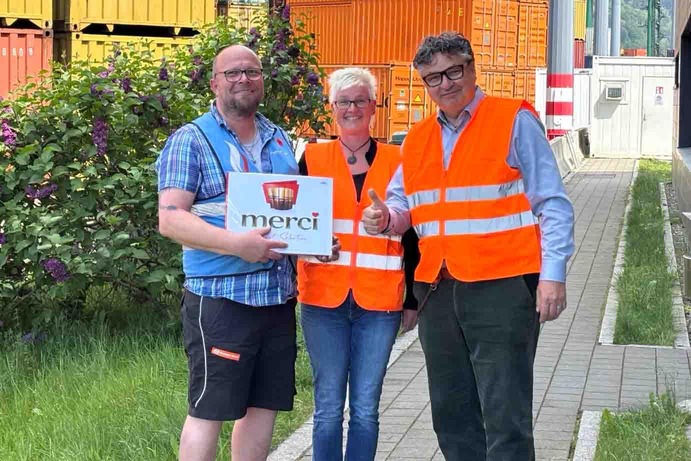 Three people stand on a path in front of stacked containers, wearing high-visibility orange vests. 