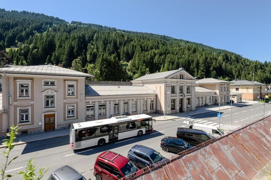 Foto Bahnhofsgebäude Bad Gastein<br/>Textinsert: Bahnhofsgebäude mit Bus und parkenden Autos