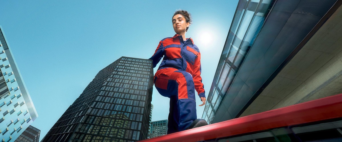 Next LevelHero Sujet der Lehrlingskampagne<br/>Textinsert: Frau mit blau-rotem Schutz-Overall balanciert auf rotem Balken, rundherum hohe Gebäude und blauer Himmel