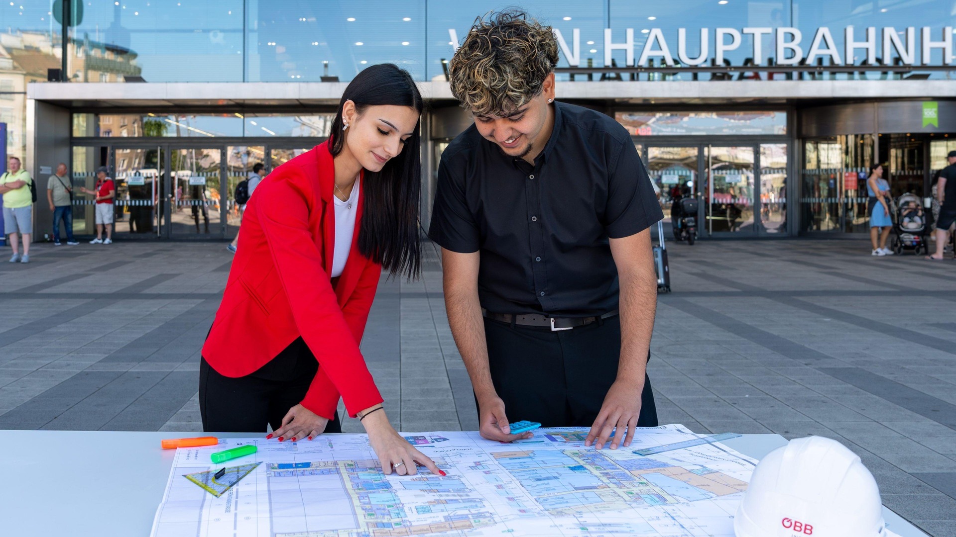 Lehrberuf Immobilienkauffrau:mann. Zwei Lehrlinge stehen vorm Hauptbahnhof Wien und schauen sich die Pläne an.