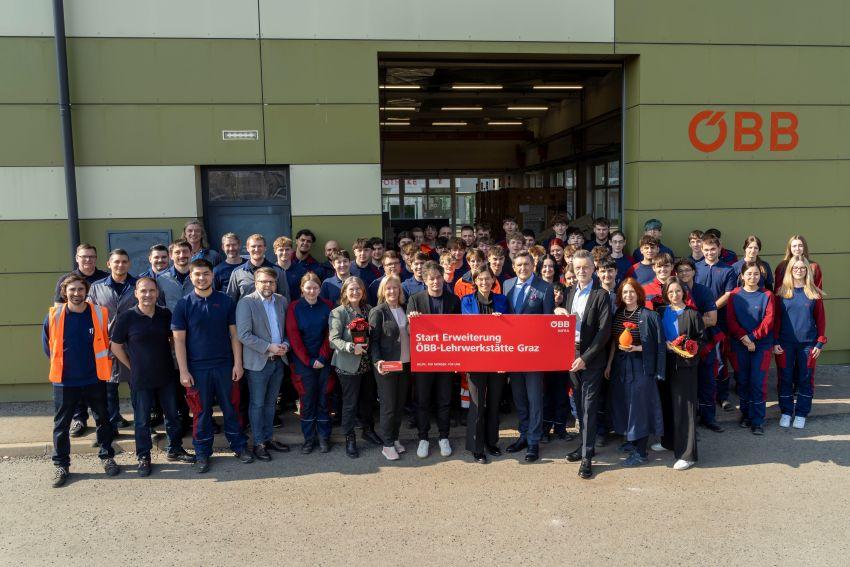 Lehrlinge stehen gemeinesam mit INFRA-Vorständin Angelo und Minister Hanke vor der Lehrwerkstätte Graz und halten ein Schild hoch. Textinsert vom Schild: Start Erweiterung ÖBB-Lehrwerkstätte Graz