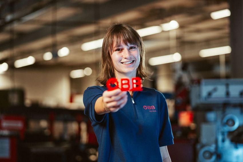Lehrling in der Lehrwerkstätte und hält ÖBB Motiv in die Kamera
