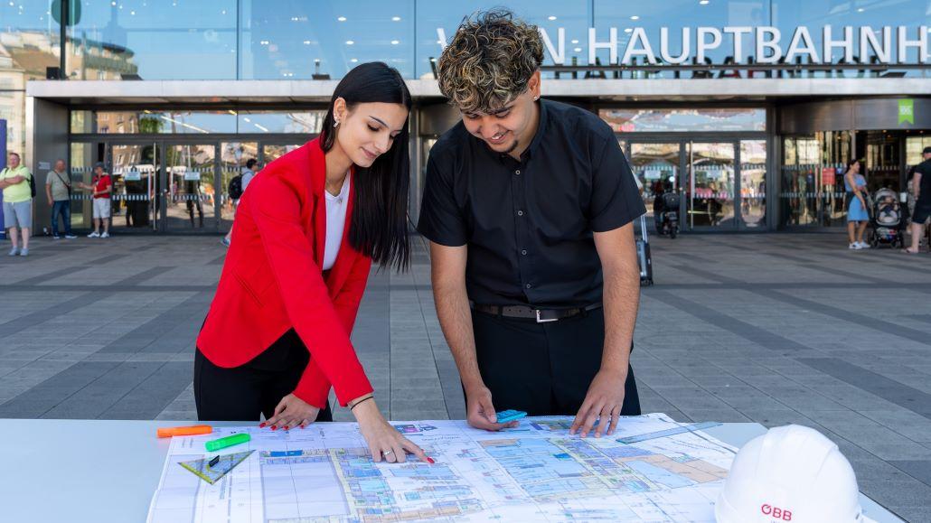 Lehrberuf Immobilienkauffrau:mann. Zwei Lehrlinge stehen vor den Hauptbahnhof Wien und schauen sie die Pläne dazu an.