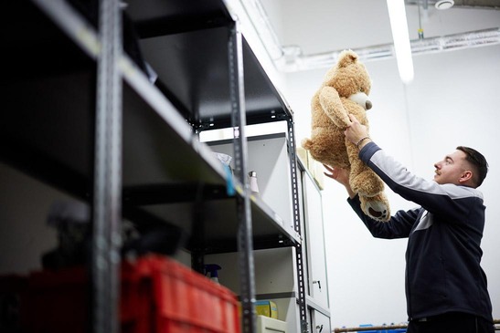 Ein Mitarbeiter des Lost and Found hebt einen großen Teddybären aus einem Regal.