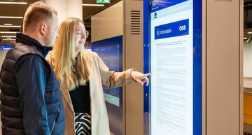 Frau und Mann stehen vor einem länglichen Monitor, die Infostele