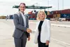 Two people are shaking hands and looking at the camera. A gantry crane and some containers can be seen in the background.