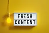 Textinsert Fresh Content auf weiß-grauem Schild, links daneben eine leuchtende Glühbirne, gelber Bildhintergrund