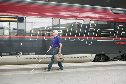 Sehbehindeter älterer Herr mit Blindenstock läuft am taktilen Leitsystem am Bahnsteig entlang. In der linken Hand hält er einen Koffer. Im Hintergrund steht ein Railjet.