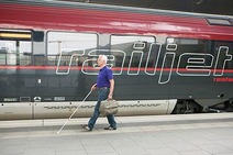 Sehbehindeter älterer Herr mit Blindenstock läuft am taktilen Leitsystem am Bahnsteig entlang. In der linken Hand hält er einen Koffer. Im Hintergrund steht ein Railjet.