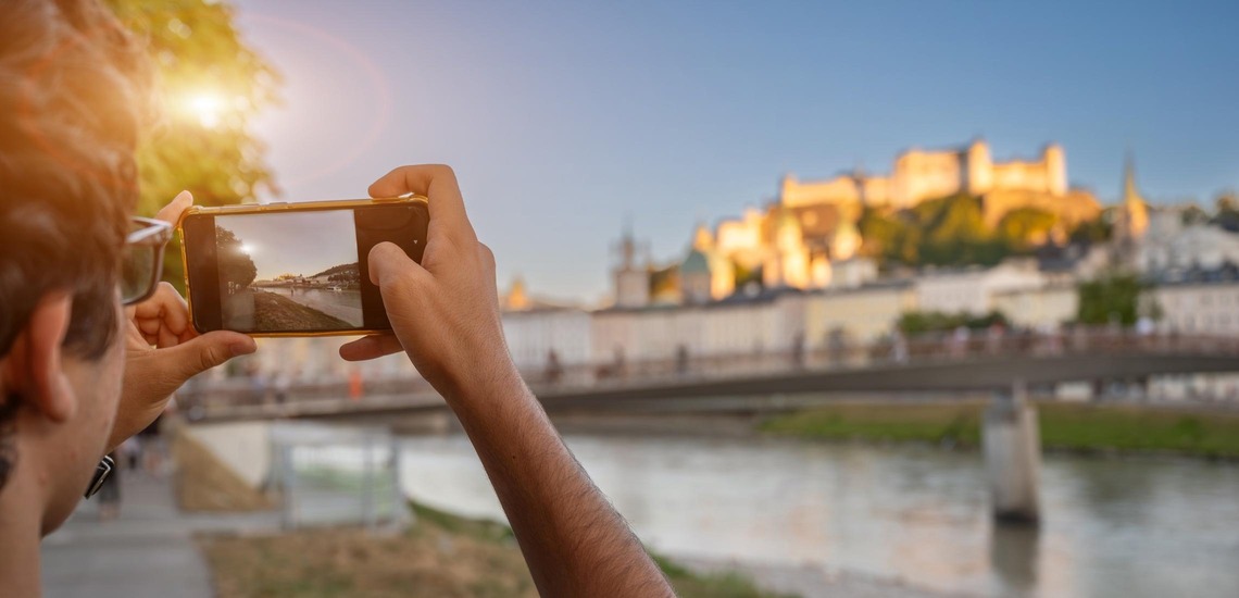 Salzburg Geheimtipps: Ein junger Mann steht am Ufer der Salzach und fotografiert mit dem Smartphone die Salzburger Festung