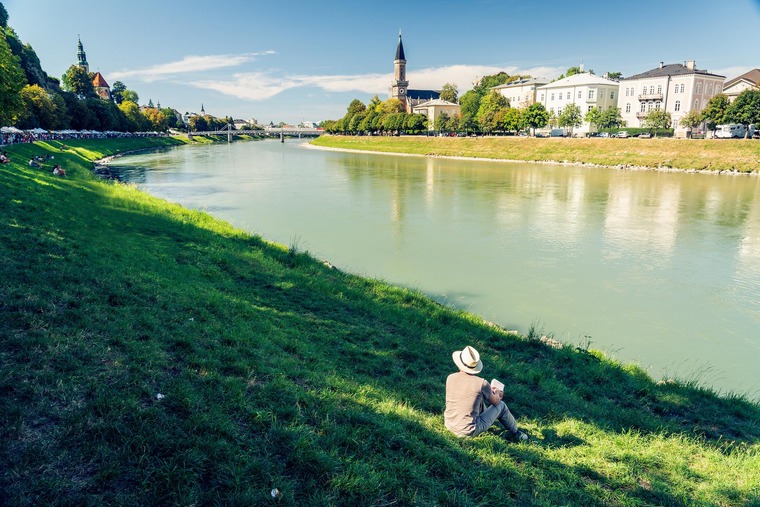 Das Salzach-Ufer gehört zu Salzburg Geheimtipps: Ein junger Mann sitzt auf dem Ufer der Salzach und blickt auf die Altstadt und die Festung auf der gegenüberliegenden Uferseite. 