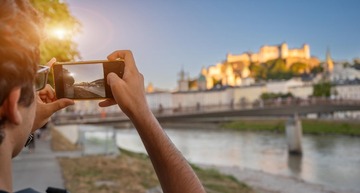Salzburg Geheimtipps: Ein junger Mann steht am Ufer der Salzach und fotografiert mit dem Smartphone die Salzburger Festung.