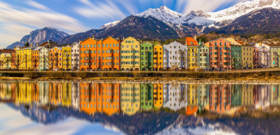 Sehenswürdigkeiten in Innsbruck: Die bunte Häuserzeile von Innsbruck, davor der Inn und dahinter die noch leicht schneebedeckten Berge Tirols. 