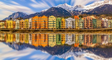 Sehenswürdigkeiten in Innsbruck: Die bunte Häuserzeile von Innsbruck, davor der Inn und dahinter die noch leicht schneebedeckten Berge Tirols. 