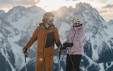 2 Personen stehen auf der Piste und sehen sich an, Hintergrund die Berge