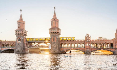 Eine junge Frau und ein Mann fahren bei untergehender Sonne vor der Oberbaumbrücke Stand-up-Paddle auf der Spree.
