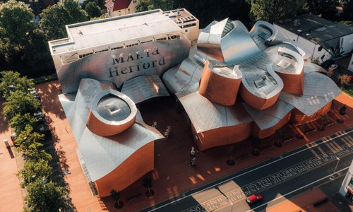Eine Luftaufnahme zeigt das Marta Herford Museum für zeitgenössische Kunst in Herford. Das Gebäude weist komplexe architektonische Formen mit einem Metalldach und roten Wandstrukturen auf. Bäume säumen den Weg auf einer Seite, und auf der Straße fährt ein rotes Auto vorbei.