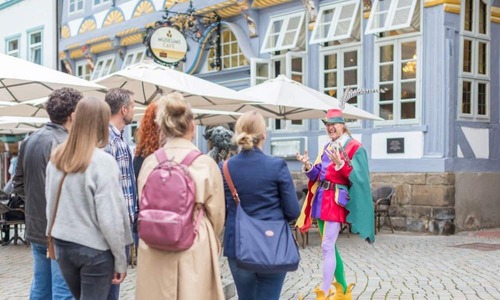 Geführte Stadtführung auf einem gepflasterten Platz vor historischen Fachwerkhäusern; eine kostümierte Figur in bunter Kleidung steht im Mittelpunkt und spricht zu einer kleinen Gruppe von Teilnehmenden, im Hintergrund sind Café‑Tische und Sonnenschirme zu sehen.