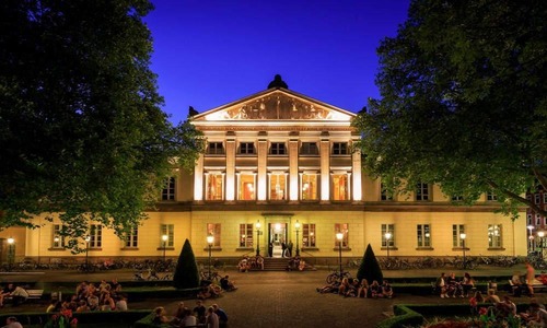 Wilhelmsplatz in Göttingen bei Nacht mit der beleuchteten Aula der Universität, umgeben von Bäumen und Menschen, die sich auf dem Platz aufhalten.