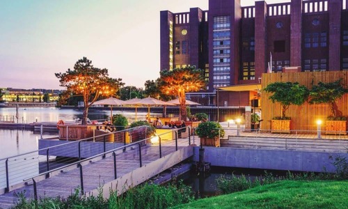 Abendstimmung in der Autostadt Wolfsburg mit beleuchteten Gebäuden, Wasserflächen und einer modernen Promenade mit Sitzbereichen und Gastronomie.