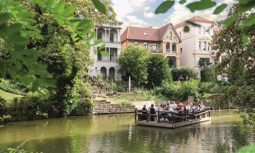 Floßfahrt auf der Oker in Braunschweig mit einer Gruppe von Menschen auf einem bewirteten Floß, umgeben von grüner Uferlandschaft und historischen Villen.