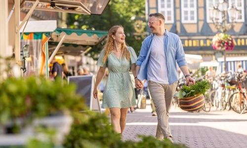 Paar schlendert über einen belebten Wochenmarkt in der Altstadt von Celle, mit frischen Einkäufen in der Hand, umgeben von Fachwerkhäusern, Marktständen und Fahrrädern.