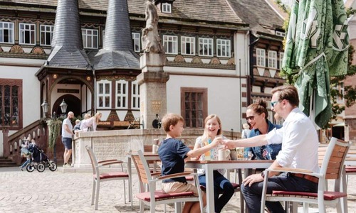 Familie sitzt in einem Straßencafé in der Altstadt von Einbeck, stößt gemeinsam an und genießt die Atmosphäre vor dem historischen Rathaus und einem Brunnen.