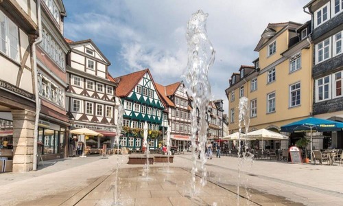 Belebter Stadtplatz in Wolfenbüttel mit Wasserspielen, umgeben von historischen Fachwerkhäusern, Geschäften und Außengastronomie.