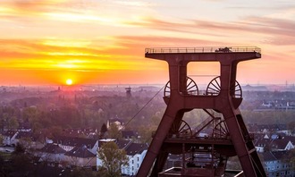 Vor dem Hintergrund der aufgehenden Sonne in Essen erhebt sich der Doppelbock-Förderturm der Zeche Zollverein in Essen. Darunter erstrecken sich Häuserzeilen und grüne Bäume. Der Himmel ist orange und gelb gefärbt.<br/>