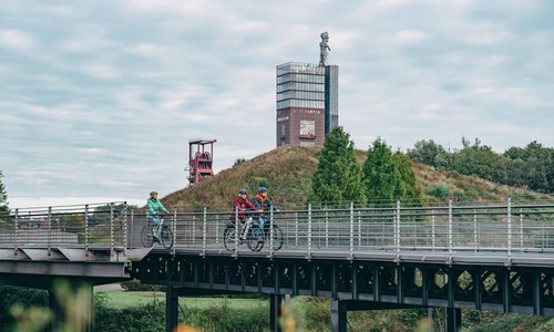 Mehrere Personen fahren mit Fahrrädern über eine erhöhte Stahlbrücke; im Hintergrund erhebt sich eine ehemalige Industrieanlage mit Turm und Fördergerüst auf einer begrünten Halde, umgeben von Bäumen unter bewölktem Himmel.<br/>