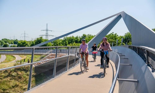 Mehrere Personen fahren mit Fahrrädern über eine moderne Fuß‑ und Radbrücke mit geschwungenen Metallstreben; seitlich verlaufen Geländer, im Hintergrund sind Gleise, Grünflächen und Strommasten unter klarem Himmel zu sehen.<br/>