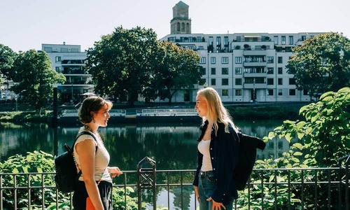 Zwei Personen stehen an einem Geländer am Ufer eines ruhigen Flusses; im Hintergrund spiegeln sich moderne Wohngebäude und Bäume im Wasser, darüber erhebt sich ein markanter Turm vor klarem Himmel.<br/>
