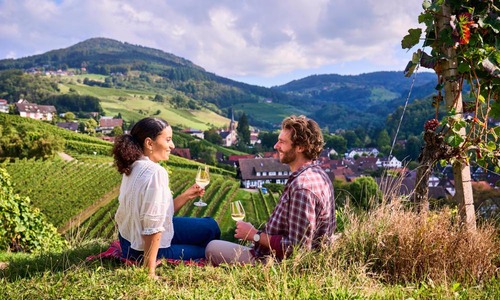 Ein Paar sitzt inmitten der Weinberge von Sasbachwalden und genießt ein Glas Wein mit Blick auf das malerische Dorf und die umliegenden Hügel