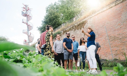 Stadtführerin erklärt einer Besuchergruppe vor hoher Ziegelmauer, daneben moderner Aussichtsturm mit offener Wendeltreppe<br/><br/>