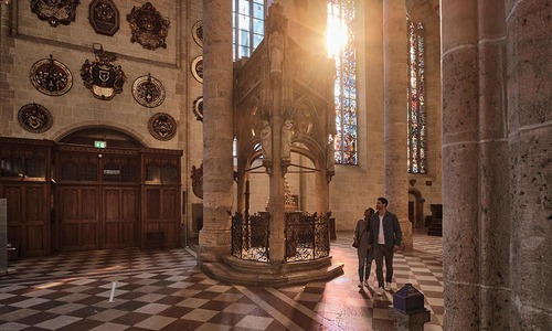 In einer Kirche mit hohen Säulen und bunten Fenstern betrachtet ein Paar ein reich verziertes steinernes Chorgestühl<br/>