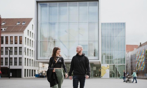 Zwei Personen gehen über einen Platz vor einem modernen Glasgebäude in der Innenstadt, Familie mit Kinderwagen im Hintergrund.<br/>