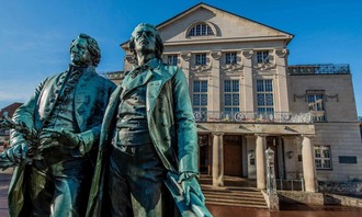 Bronzeskulptur zweier Figuren im Vordergrund, dahinter ein klassizistisches Theatergebäude mit Säulenfassade und breiter Freitreppe.<br/>