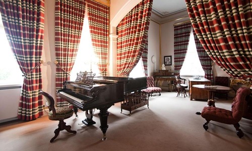 Eleganter historischer Salon mit schwarzem Flügel, hohen Fenstern und schweren, rot gemusterten Vorhängen; klassische Möbel schaffen eine repräsentative Atmosphäre.<br/>