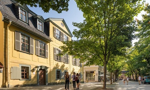Gelbes historisches Wohnhaus an einer baumbestandenen Straße; Menschen flanieren auf dem Gehweg vor der klassizistischen Fassade.<br/>