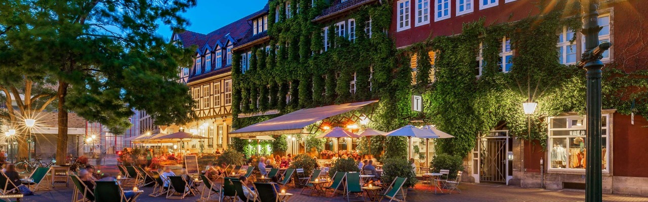 Belebter Ballhofplatz in Hannover am Abend mit Außengastronomie, Menschen auf Liegestühlen und Tischen, warmem Licht sowie historischen, teils begrünten Fachwerkhäusern im Hintergrund.