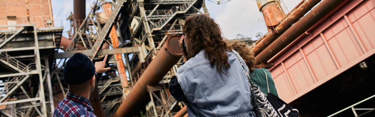 Mehrere Personen stehen vor einer großflächigen historischen Industrieanlage mit Stahlgerüsten, Rohrleitungen und Hochöfen; eine Person zeigt auf die komplexe Konstruktion, die sich vor hellem Himmel erhebt.<br/>