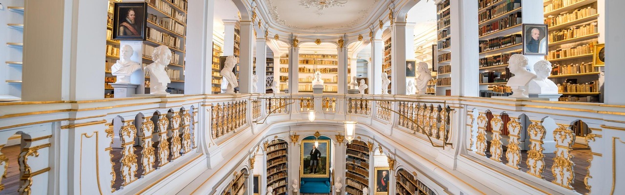 Prunkvoller Bibliothekssaal mit umlaufenden Galerien, goldverzierten Balustraden, weißen Büsten und deckenhohen Bücherregalen.<br/>