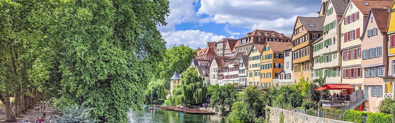 Fluss mit Stocherkähnen im Sommer, an den Ufern bunte Altstadthäuser und ein baumbestandener Fußweg.